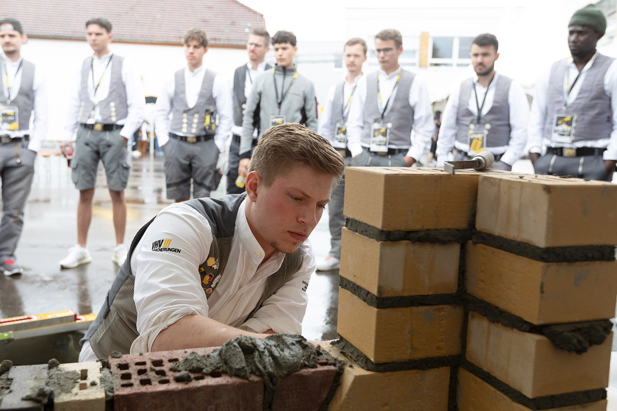 Pierre Holze, Maurerweltmeister bei den WorldSkills 2022, errichtet eine Mauer mit Annweilerer Wappen inklusive Wasserwaage und steht den Auszubildenden Rede und Antwort.